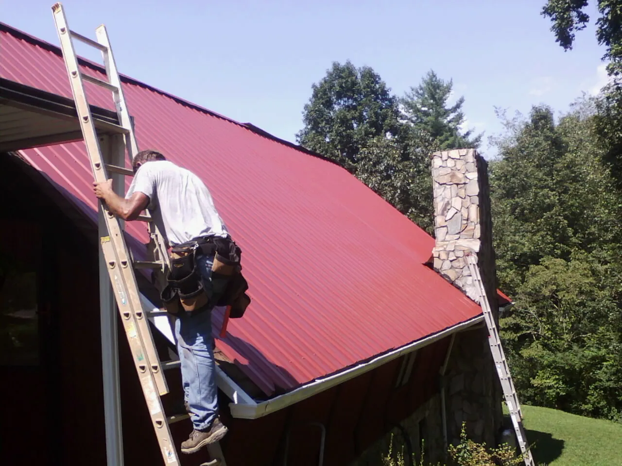 Roofer on a ladder beside a red metal roof and stone chimney — Soffit & Fascia Repair by Roofing Brothers in Schenectady